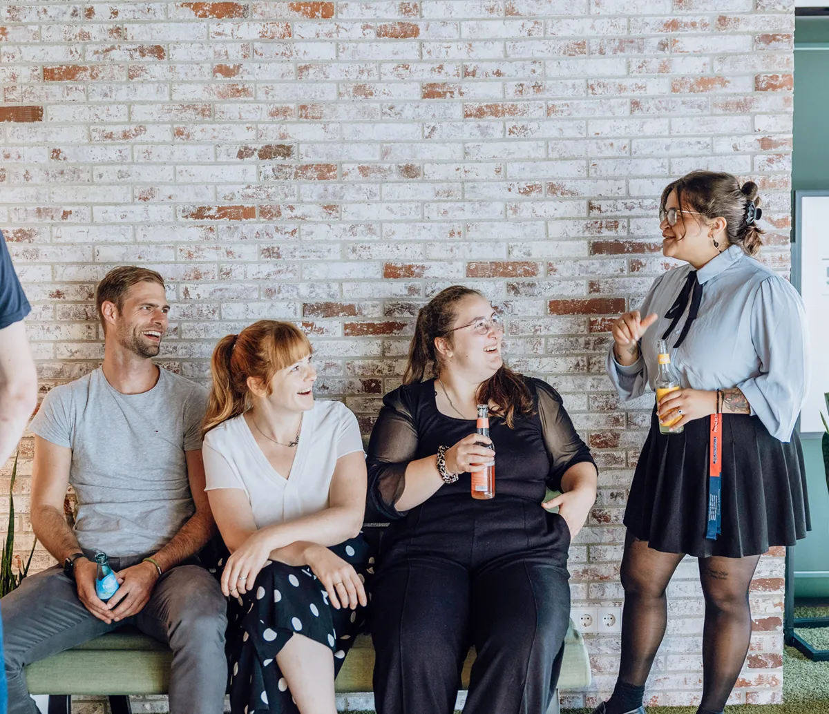 A group of people sitting and standing against a brick wall, smiling and holding drinks, engaged in a casual conversation.