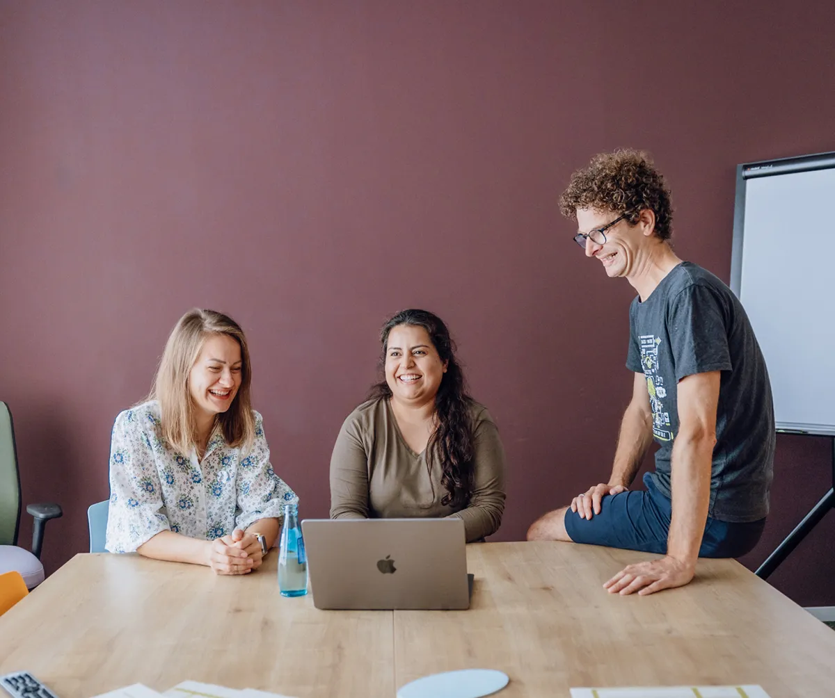 Three people smiling and sitting around a table with a laptop, in a casual office setting.