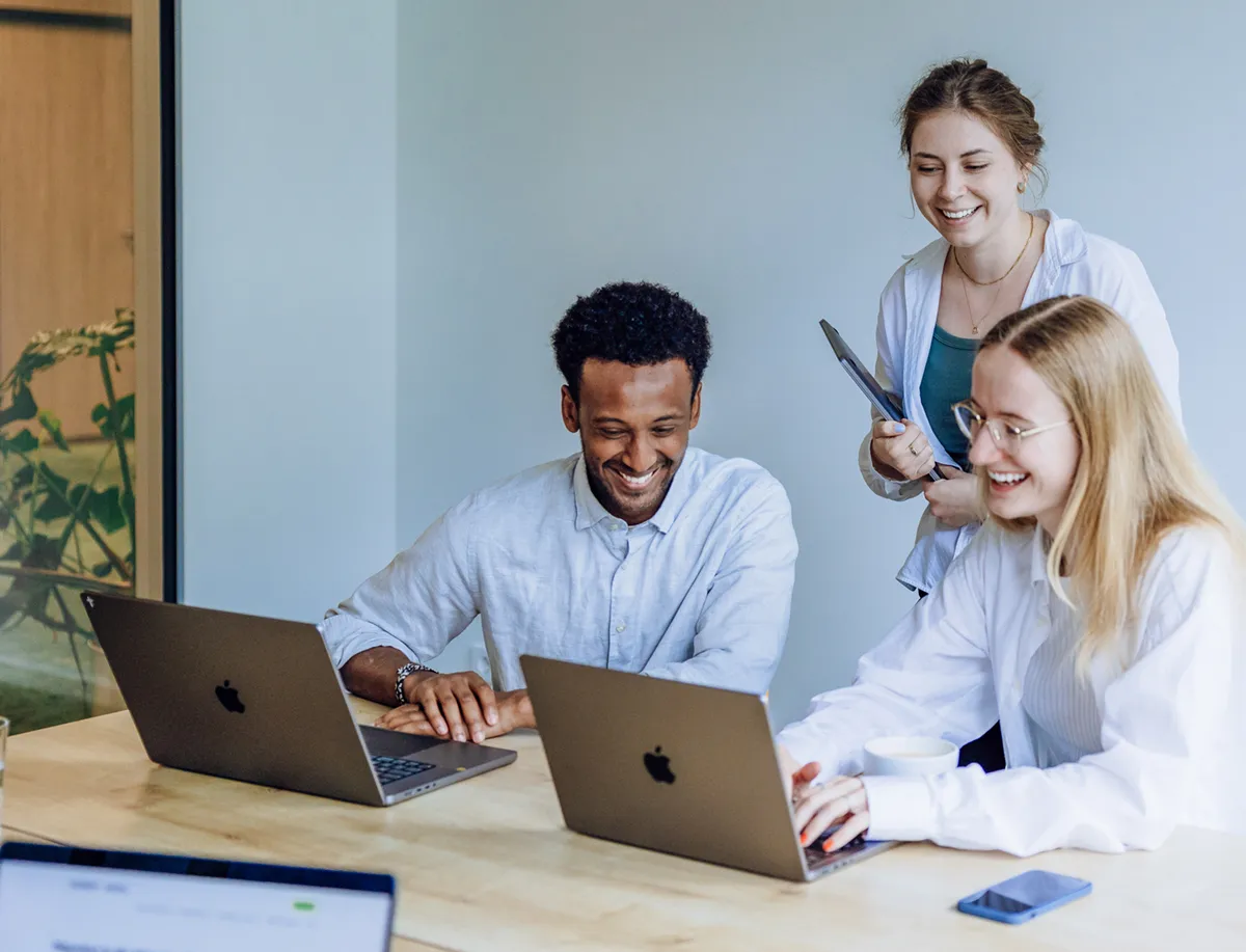 Three colleagues smiling and working together on laptops at a wooden table in a bright office space.