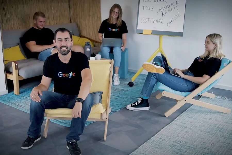 Four colleagues wearing black Google t-shirts relax in a casual workspace featuring a rug, lounge chairs, and a whiteboard with the words "Teamwork," "Tools," "Software," and "Zusammenarbeit" written on it.