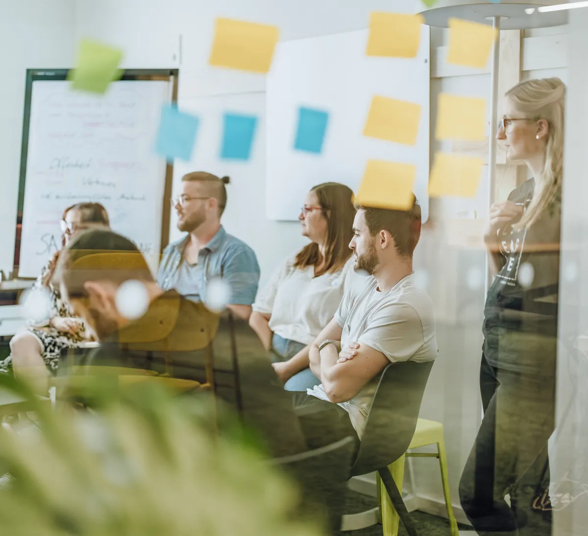 A group of people sitting and standing in a meeting room, viewed through a glass wall with sticky notes attached.