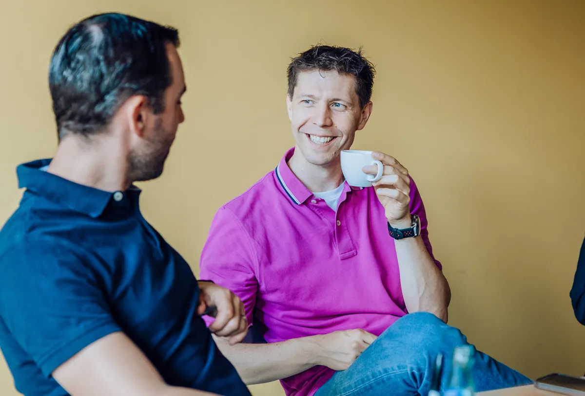 Two men sitting and smiling while having a conversation, one holding a coffee cup, against a yellow background.
