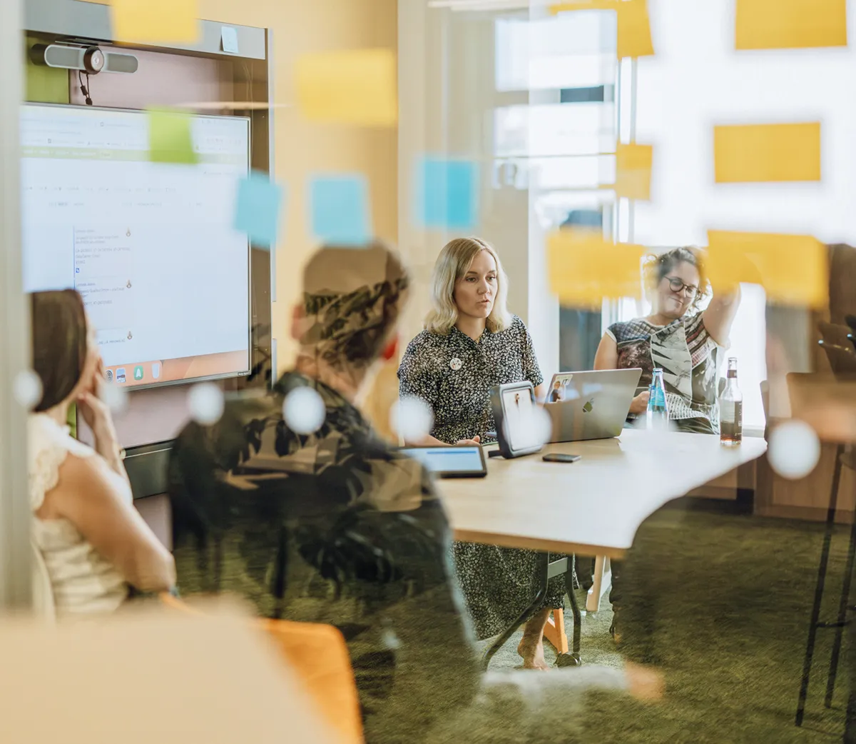 Office meeting with people sitting around a table, visible through a glass wall with sticky notes.